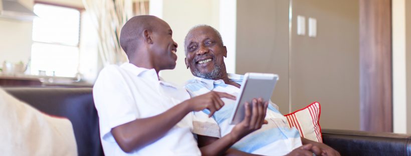 Picture of an older man and a younger man on a couch looking at a tablet screen laughing.