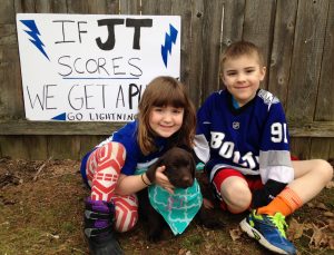 Picture of two kids sitting on the ground next to a chocolate lab puppy.